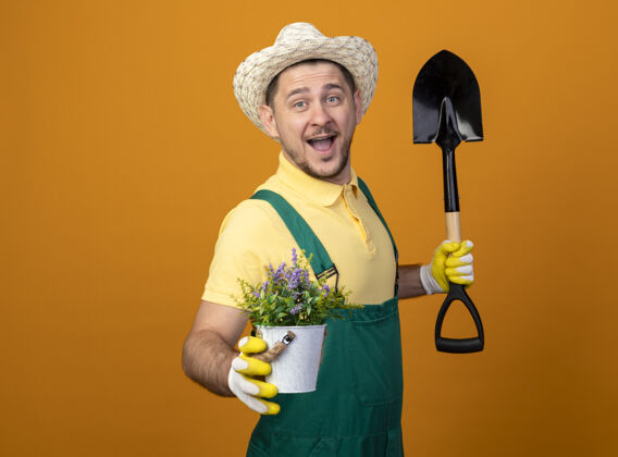 连身衣年轻的园丁穿着连体衣 戴着帽子 拿着铲子 愉快地微笑着展示盆栽植物帽子橘子微笑