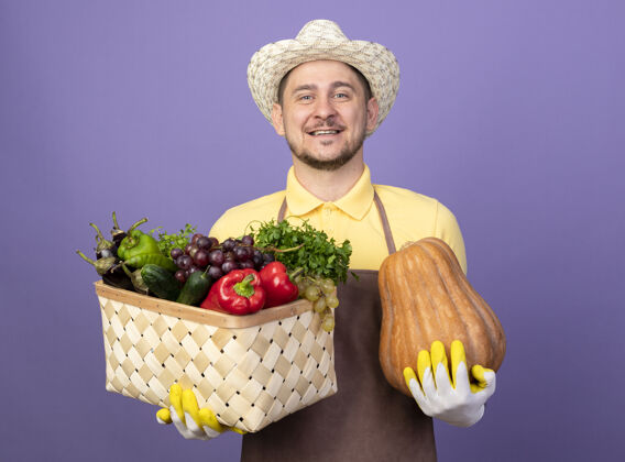 满的年轻的园丁 穿着连体衣 戴着帽子 戴着工作手套 手里拿着装满蔬菜和南瓜的箱子 微笑着 快乐而积极积极看抱着