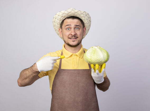 花园一个年轻的园丁 穿着连体衣 戴着帽子 戴着工作手套 手里拿着卷心菜 指挥员微笑着指着卷心菜站着微笑连身衣