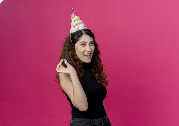 卷发年轻漂亮的女士 卷发 戴着节日帽 手持口哨 站在粉色的墙上 快乐而积极的生日派对理念站着年轻女人