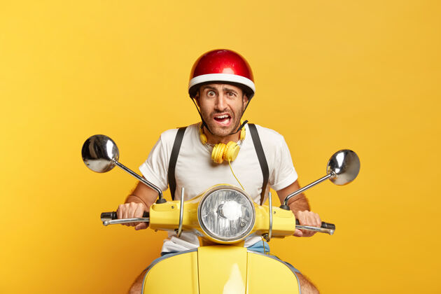 姿势不高兴的英俊男司机骑着红色头盔的滑板车乘坐耳机运输
