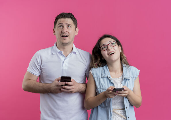 移动一对穿着休闲服的年轻夫妇举着智能手机 站在粉色的墙上 神色恍惚地仰望着年轻衣服抱