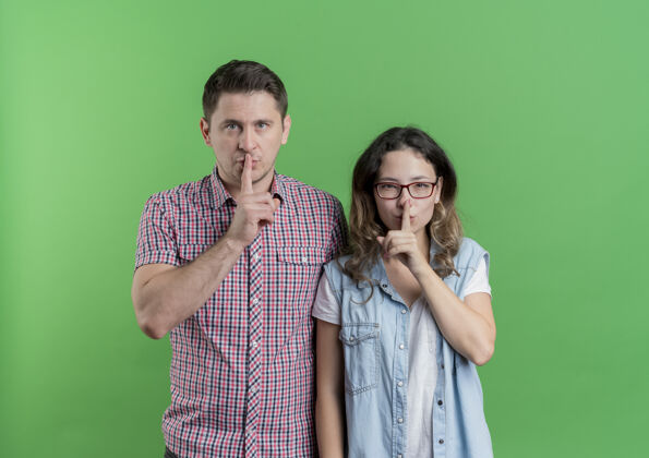 女人一对穿着休闲服的年轻夫妇站在绿色的墙上 手指放在嘴唇上做着沉默的手势站着情侣手势