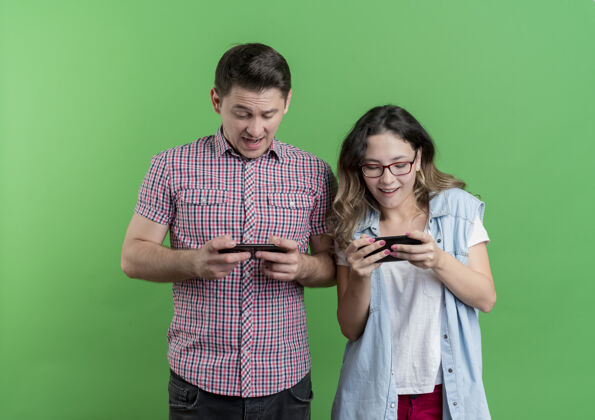 年轻一对穿着休闲服的年轻夫妇手持智能手机 站在绿墙上玩游戏站着游戏休闲