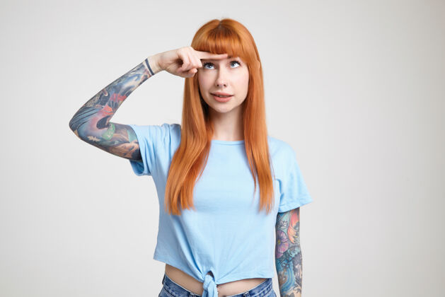 长迷惑不解的年轻漂亮的纹身女子 留着一头蓬松的长发 把食指放在额头上 一边看着它 一边穿着蓝色t恤站在白色背景上表情白皙食指