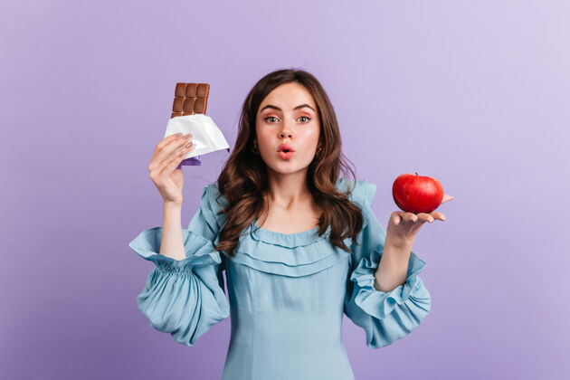 欢乐穿蓝色上衣的女人在紫色的墙上摆姿势漂亮的女孩在思考她的饮食 在多汁的苹果和脂肪巧克力之间做出选择苹果魅力卷发