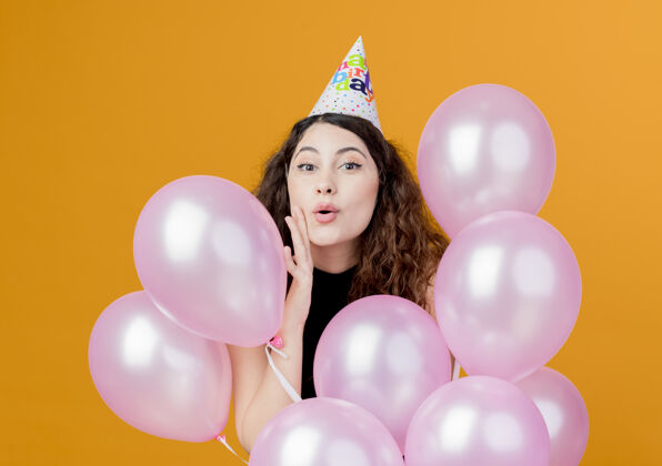 气球一位年轻漂亮的女士 卷发 戴着节日帽 手持气球 站在橙色的墙上 快乐而兴奋地庆祝生日空气抱着站着