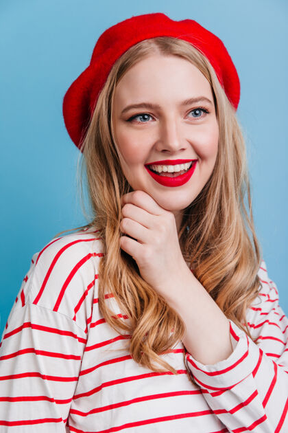 优雅表现积极情绪的金发美女穿法国贝雷帽的时髦女孩在蓝色的墙上化妆快乐发型