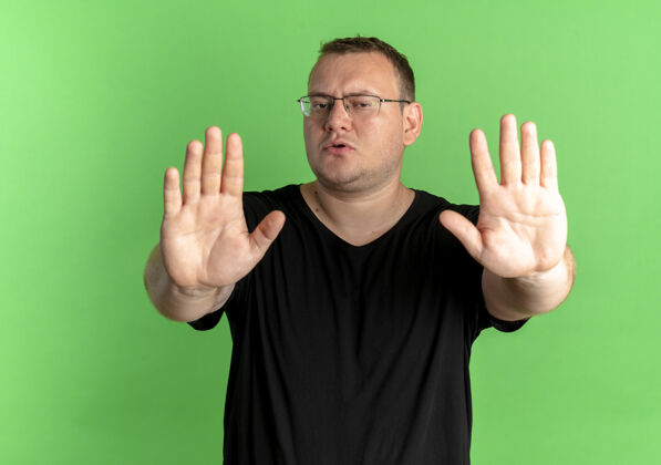 男人戴眼镜的超重男子身穿黑色t恤 双手张开站在绿色墙壁上停止唱歌制造戴歌手