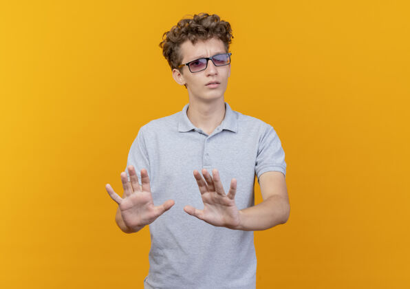 眼镜一个戴着黑眼镜 穿着灰色马球衫的年轻人站在橘色的墙上 手举着手示意不要靠近穿来年轻