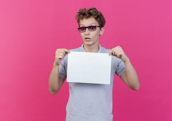 床单一个戴着黑眼镜的年轻人穿着灰色马球衫 手里拿着一张白纸 站在粉色的墙上惊讶地看着穿男人秀