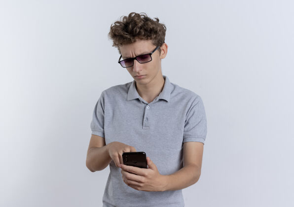 男人一个戴着黑眼镜 穿着灰色马球衫的年轻人站在白墙上 严肃地看着他的智能手机手机年轻看移动