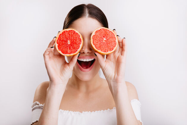 年轻表情积极的快乐女孩用橘子遮住眼睛 在孤零零的墙上摆姿势女性不化妆化妆