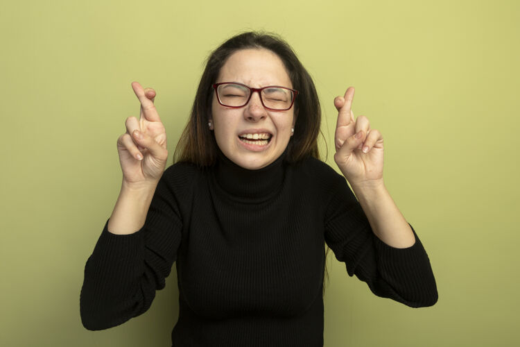 站年轻漂亮的女孩穿着黑色高领毛衣戴着眼镜许下美好的愿望闭上眼睛望着希望的表情交叉手指美丽交叉愿望