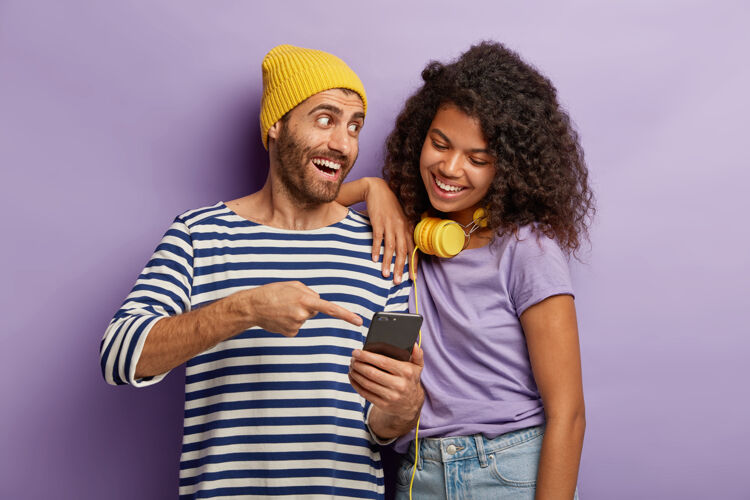 条纹戴着黄色帽子和条纹套头衫的快乐男人 向非洲女孩解释如何在智能手机上使用新应用程序 指着屏幕 站得很近 无法想象没有现代科技的生活耳机手机黄色