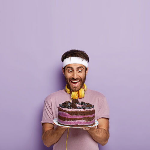 音乐喜出望外的惊喜男人盯着美味的蛋糕 感觉到诱惑 运动后饿了 穿着休闲t恤 戴着耳机高兴美味健康