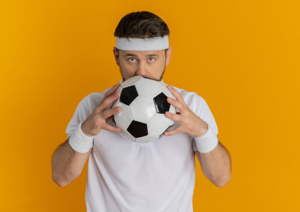 拿着身穿白衬衫的年轻健身男子 头上戴着足球带 脸藏在足球后面 站在橙色背景上站着足球健身
