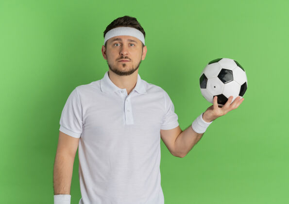 拿着身穿白衬衫 头戴足球带的年轻健身男子站在绿色背景下 严肃地看着摄像机头带男人足球