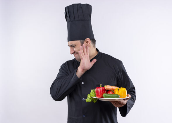 蔬菜男厨师身穿黑色制服 头戴厨师帽 拿着盛有新鲜蔬菜的盘子 站在白色背景下 带着厌恶的表情看着一旁厨师站着穿