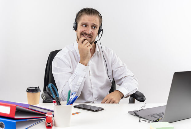 笔记本电脑穿着白衬衫 戴着耳机 拿着麦克风的帅哥坐在白色背景下的办公桌旁 紧张而紧张地工作着白色工作紧张