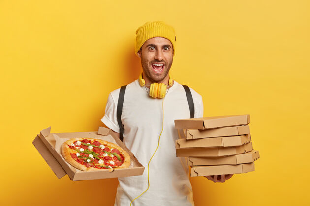快乐开朗的披萨送货员拿着纸箱站着 等着客人 戴着黄色的帽子和白色的t恤 在运送快餐的过程中听音乐快递帽子欢呼