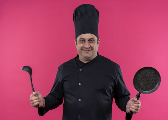 帽子男厨师身穿黑色制服 头戴厨师帽 手拿锅和勺子 自信地站在粉色背景下微笑勺子黑色微笑