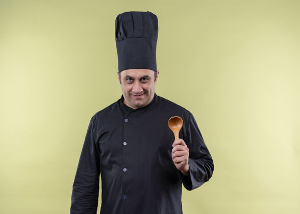 帽子男厨师身穿黑色制服 头戴厨师帽 露出木勺 站在绿色背景下 面带微笑地看着摄像机勺子绿色穿