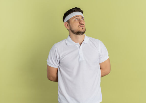 站身穿白衬衫 头箍 困惑地站在橄榄色背景下的年轻健身男子头带男人健身