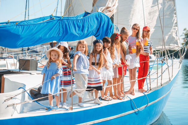女孩孩子们在游艇上喝着橙汁青少年或少女们在蓝天下户外五颜六色的衣服孩子们的时尚 阳光明媚的夏天 河流和假日的概念帆船交通旅行