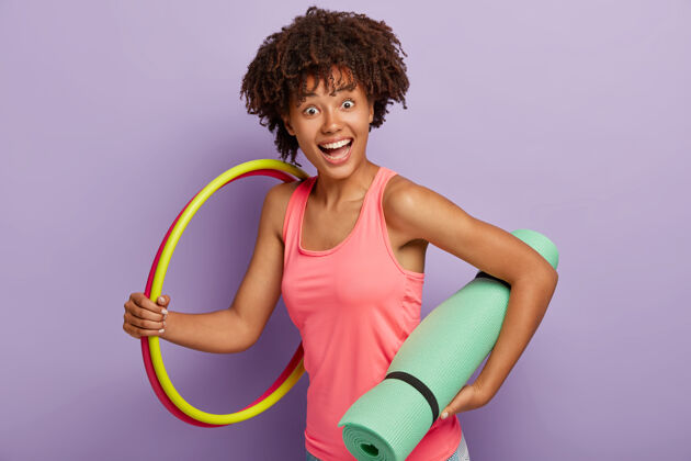 运动快乐的黑皮肤女孩背着绿松石垫子 两个篮球 在家庭氛围中锻炼身体 为了健康而从事体育运动 站在室内靠着紫墙运动理念运动装积极欢呼