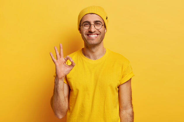 微笑帅哥的肖像做了一个很好的手势 表示同意 喜欢想法 开心地微笑 戴着眼镜 戴着黄色帽子和t恤 在室内做模特很好 谢谢手势休闲人好
