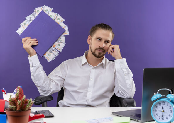 白色穿着白衬衫的帅哥商人手里拿着一个夹着现金的文件夹 带着沉思的表情坐在紫色背景的办公桌旁坐着衬衫桌子
