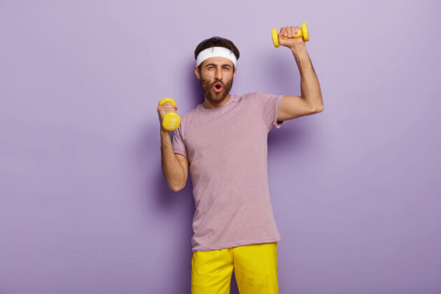 情绪有趣的男人有乐趣 练习哑铃 穿着运动服 为健康的生活方式而努力 早上有规律的训练紫色有趣头带