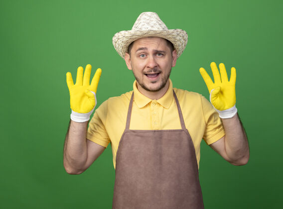 工人年轻的园丁穿着连体衣 戴着帽子 戴着工作手套 站在绿色的墙边 手指着八号微笑着穿手指站立