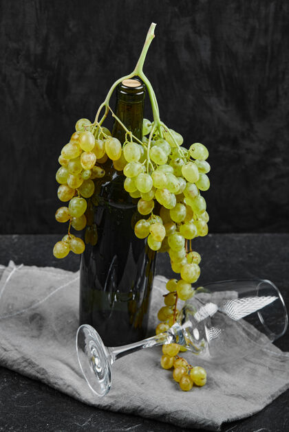 瓶子白葡萄围绕着一瓶葡萄酒和一个空杯子在黑暗的表面上用灰色桌布自然的串葡萄