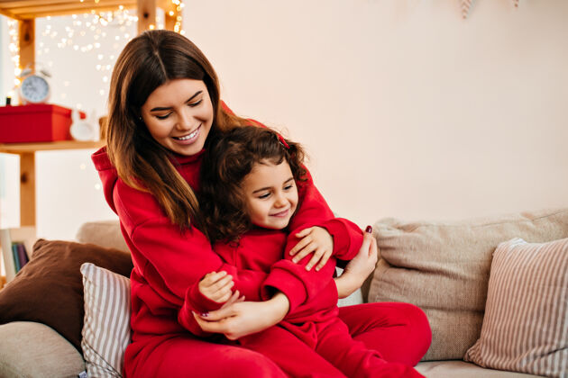 享受漂亮的黑发女人微笑着抱着孩子穿着红色衣服的年轻妈妈和小女儿坐在沙发上卷发拥抱寒冷