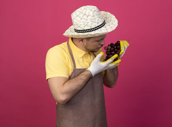 花园年轻的园丁穿着连体衣 戴着帽子 戴着工作手套 手里拿着一束葡萄 站在粉红色的墙上严肃地看着它严肃穿站着