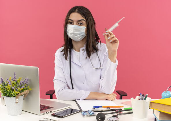 面部年轻的女医生穿着白大褂 戴着面罩 脖子上戴着听诊器 手里拿着注射器坐在桌边 笔记本电脑放在粉色的墙上工作抱着办公室