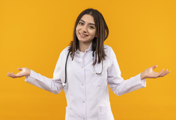 外套年轻的女医生穿着白大褂 脖子上戴着听诊器 困惑地看着前方 毫无顾忌地张开双臂站在橙色的墙上站看医生