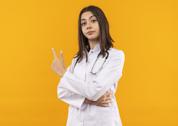 人年轻的女医生穿着白大褂 脖子上戴着听诊器 食指指向一边 站在橘色的墙上 看上去很自信外套听诊器站