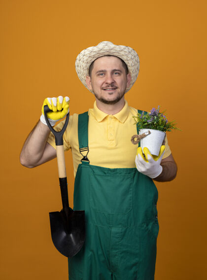 铲子年轻的园丁穿着连体衣 戴着帽子 拿着铲子和盆栽植物 站在橙色的墙上 面带微笑地看着前方人帽子微笑