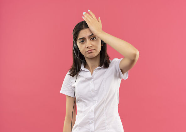 人一个穿着白衬衫戴着耳机的年轻女孩 望着前面 困惑地用手捂着头 因为她站在粉红色的墙上犯了错误麦克风站人
