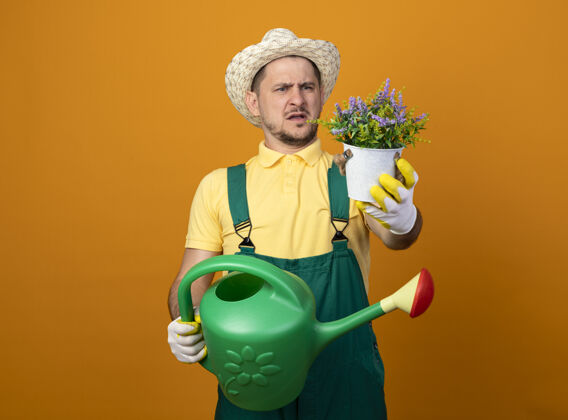 市民年轻的园丁穿着连体衣 戴着帽子 手里拿着浇水罐 站在橘红色的墙上 看着手中的盆栽植物 既不高兴又困惑站着制服植物