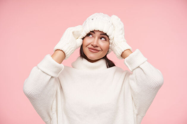 表情室内拍摄的年轻漂亮的棕色头发的女人触摸她的白色帽子 举起双手 微微微笑 穿着舒适的衣服站在粉红色的墙上女人提高自然