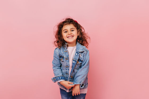 夹克快乐的卷发少年在镜头前笑无忧无虑的小女孩被隔离在粉红色背景的摄影棚拍摄积极孩子时尚