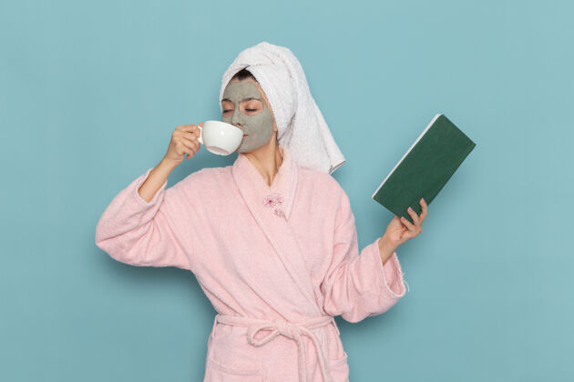年轻人正面图穿着粉色浴袍的年轻女性喝着咖啡 读着蓝墙上的文案清洁美容自理霜淋浴服装成人清洁