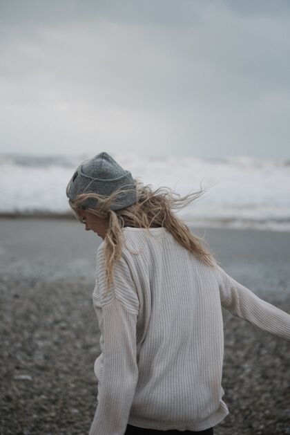 户外年轻的金发女郎戴着帽子 在多风的天气里走在多岩石的海岸上海岸海洋景点