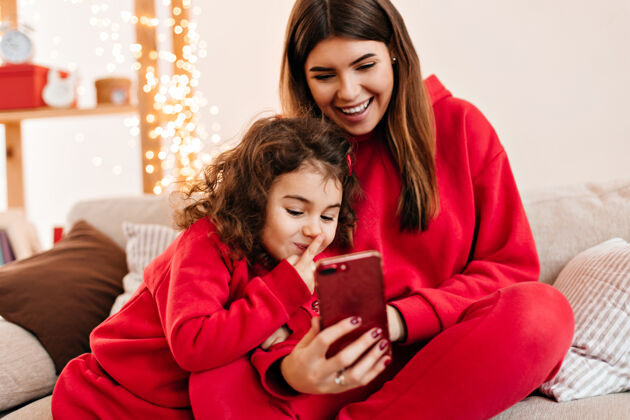 女孩可爱的卷发女孩和妈妈一起用智能手机微笑的年轻妈妈和未成年的女儿坐在沙发上女儿亲子关系深色头发
