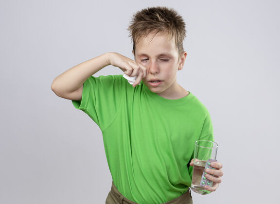 餐巾生病的小男孩穿着绿色t恤 站在白色的墙上 手里拿着一杯水和药丸 用纸巾擦鼻子 忍受着寒冷痛苦站着小