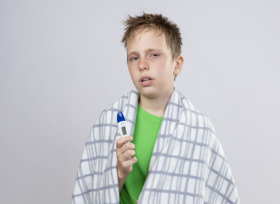 男孩生病的小男孩穿着绿色t恤 裹着毯子 拿着温度计站在白色的墙上 感觉不舒服毯子站着小